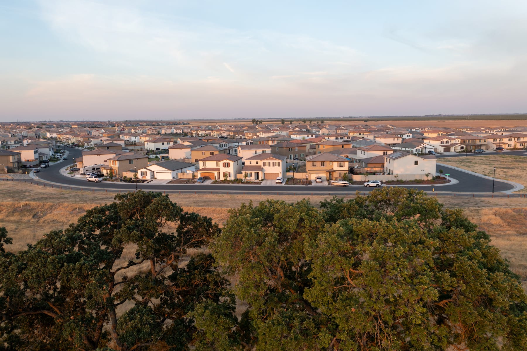 A sprawling suburban neighborhood nestled among rolling hills, with winding roads and a mix of residential homes in the foreground, set against a hazy, distant horizon.