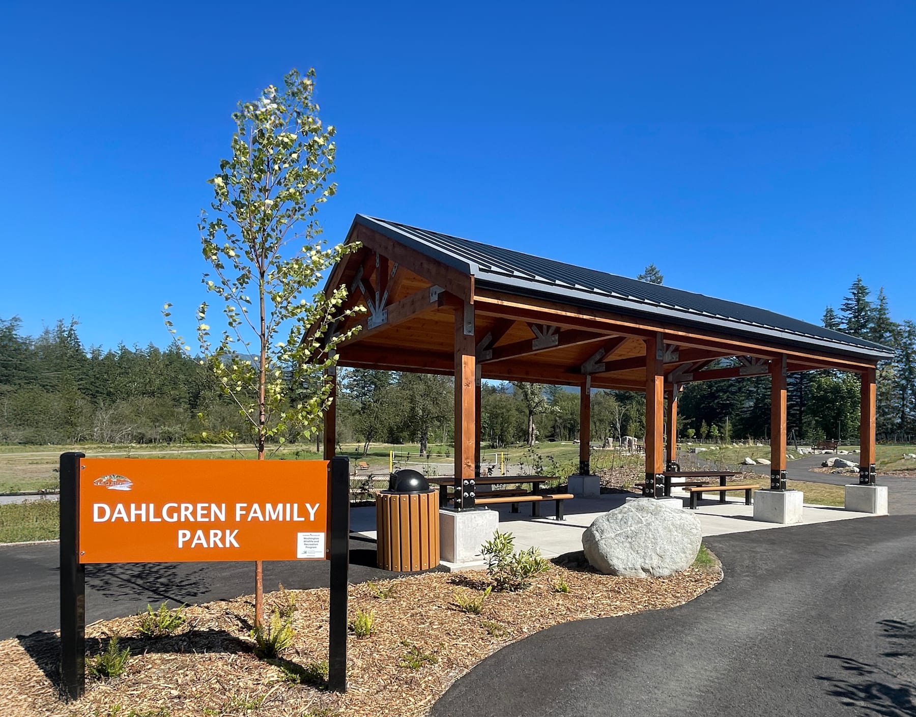 A wooden pavilion with picnic tables sits in a grassy area surrounded by trees, with a sign indicating it is the Dahlgren Family Park.