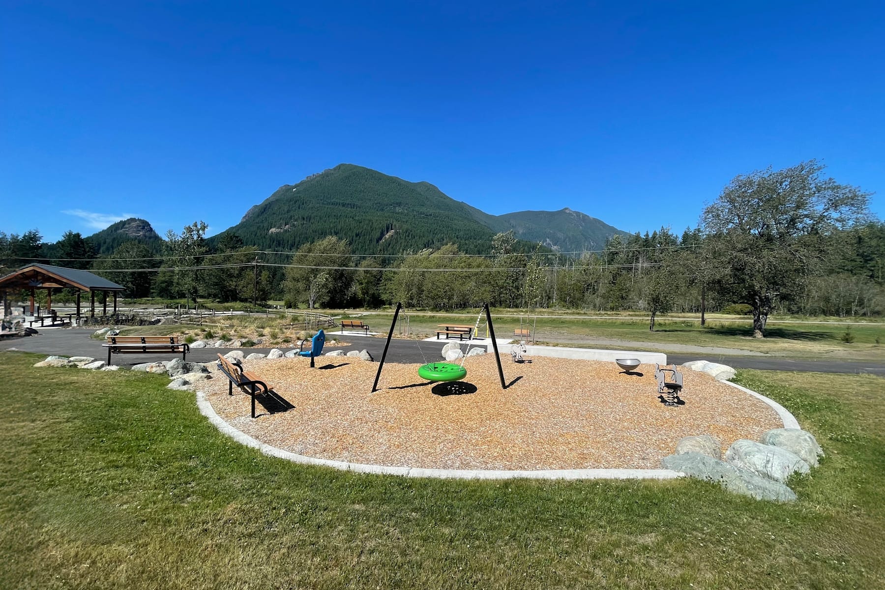 A playground with various colorful equipment sits in a grassy area, surrounded by lush trees and mountains in the background under a clear blue sky.