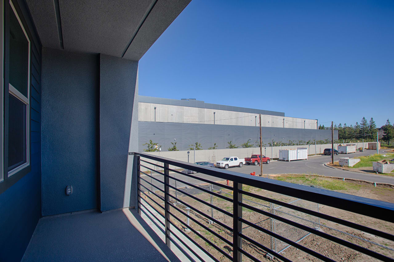 The image shows a modern industrial building with a balcony in the foreground, overlooking a parking lot and other buildings in the background under a clear blue sky.