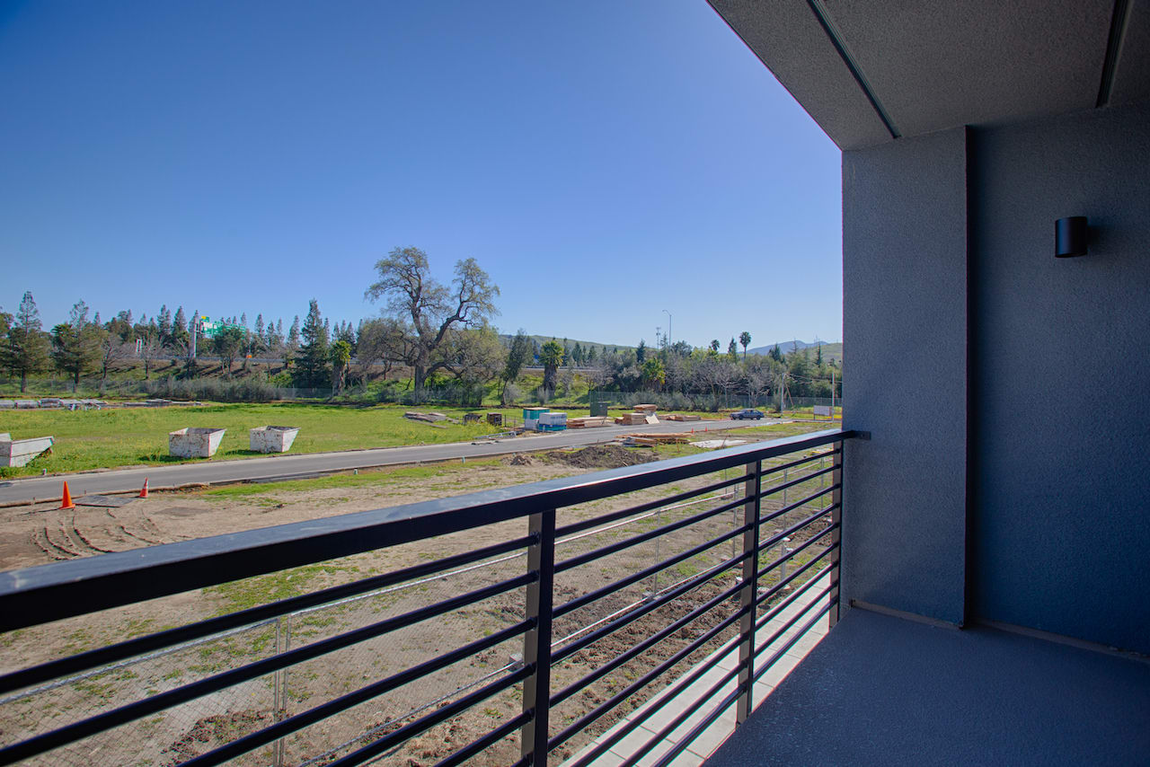 The image shows a scenic outdoor view from a balcony or porch, with a grassy field in the foreground and a wooded area with trees in the background under a clear blue sky.