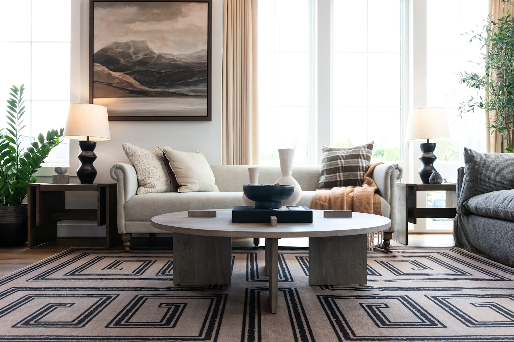 A cozy and inviting living room with a large framed landscape painting on the wall, a round coffee table with decorative items, and a plush, neutral-toned sofa complemented by patterned throw pillows.