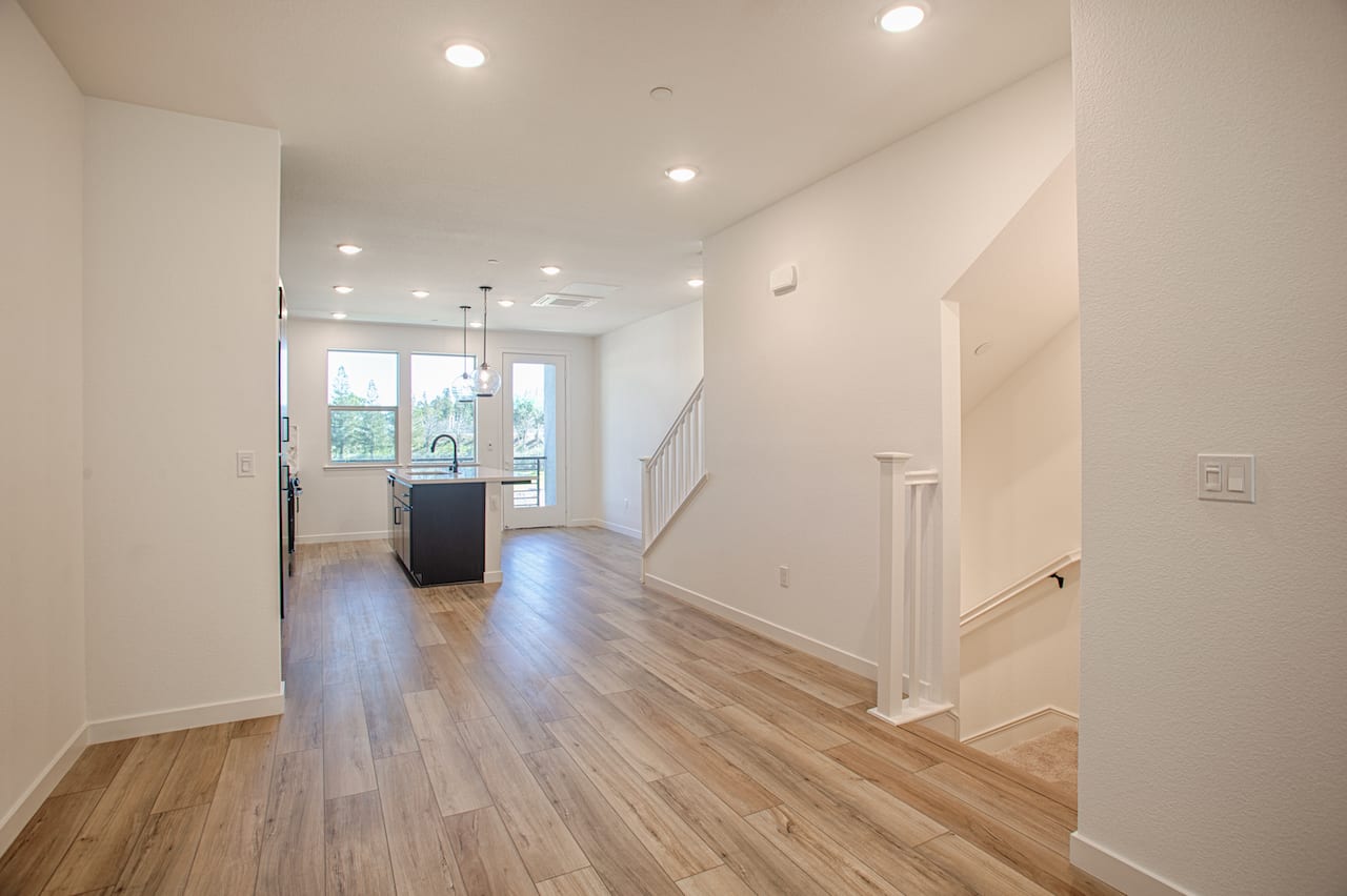 A bright and spacious hallway with hardwood floors, recessed lighting, and a staircase leading to the upper level. The hallway opens up to a kitchen area with a dark-colored cabinet and appliances visible in the background.