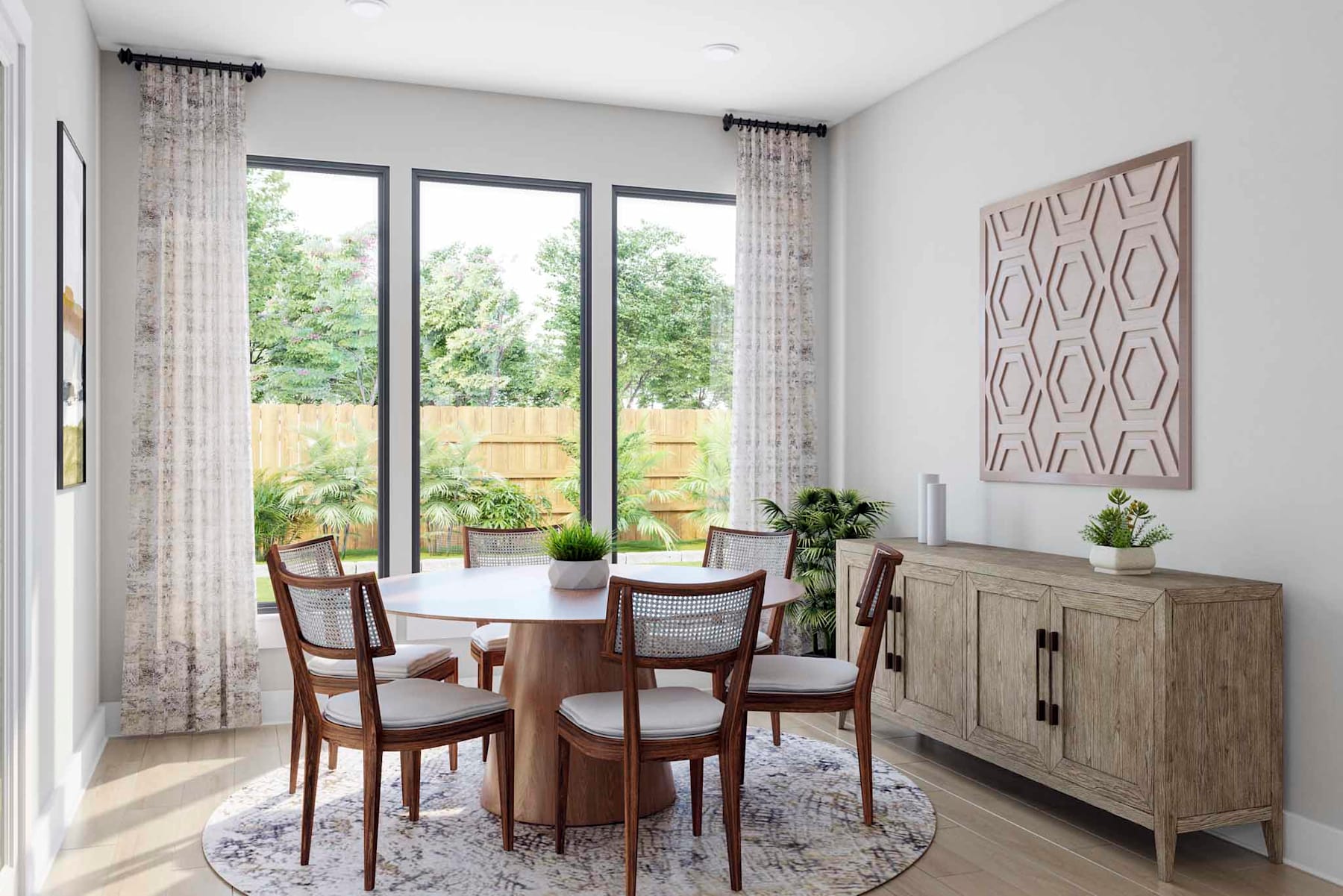 A cozy dining area with a wooden table and chairs, surrounded by large windows overlooking a lush outdoor landscape, complemented by a patterned wall art and a wooden cabinet.