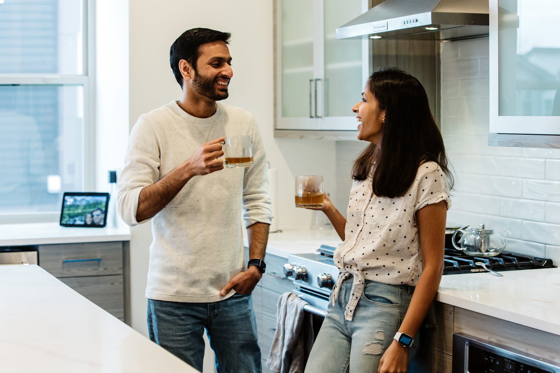 A smiling couple standing in a kitchen, engaged in conversation and holding drinks.