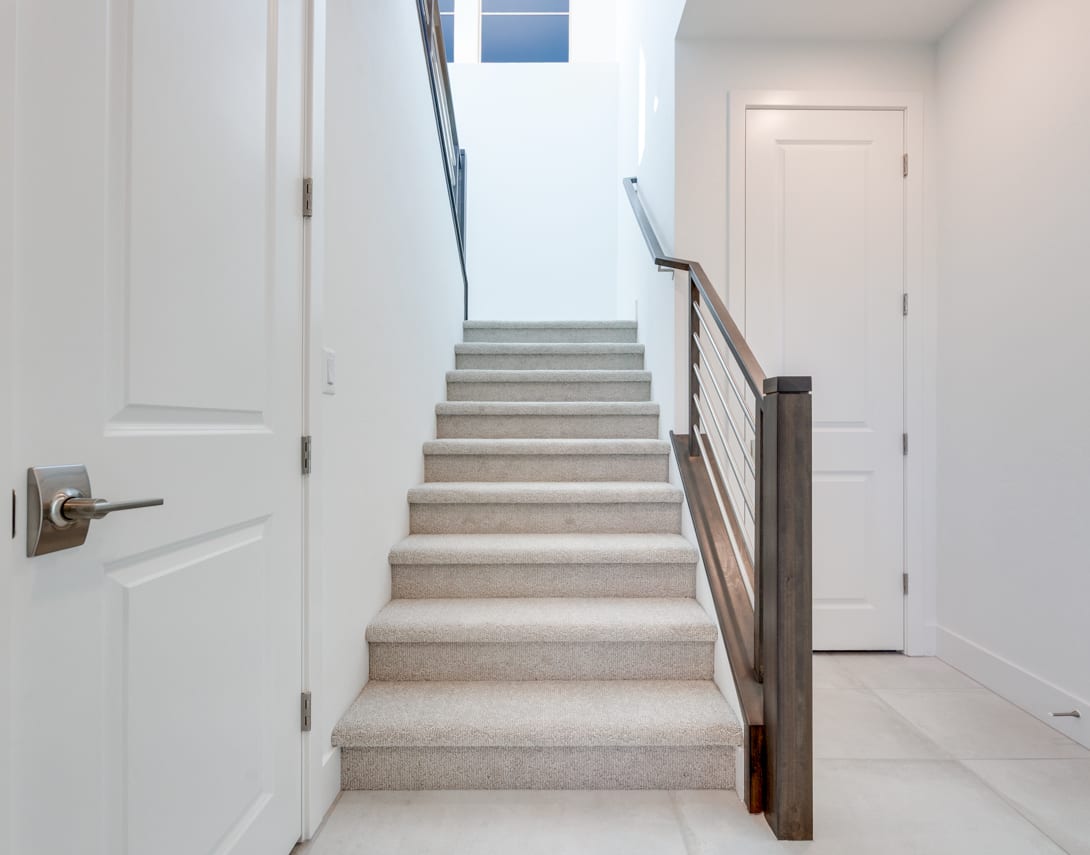 A well-lit staircase with white walls and a wooden handrail leads up to a doorway in the background.