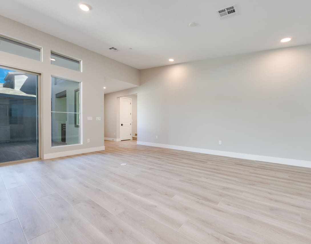 An empty, spacious room with light-colored hardwood floors, white walls, and a sliding glass door leading to an outdoor area.