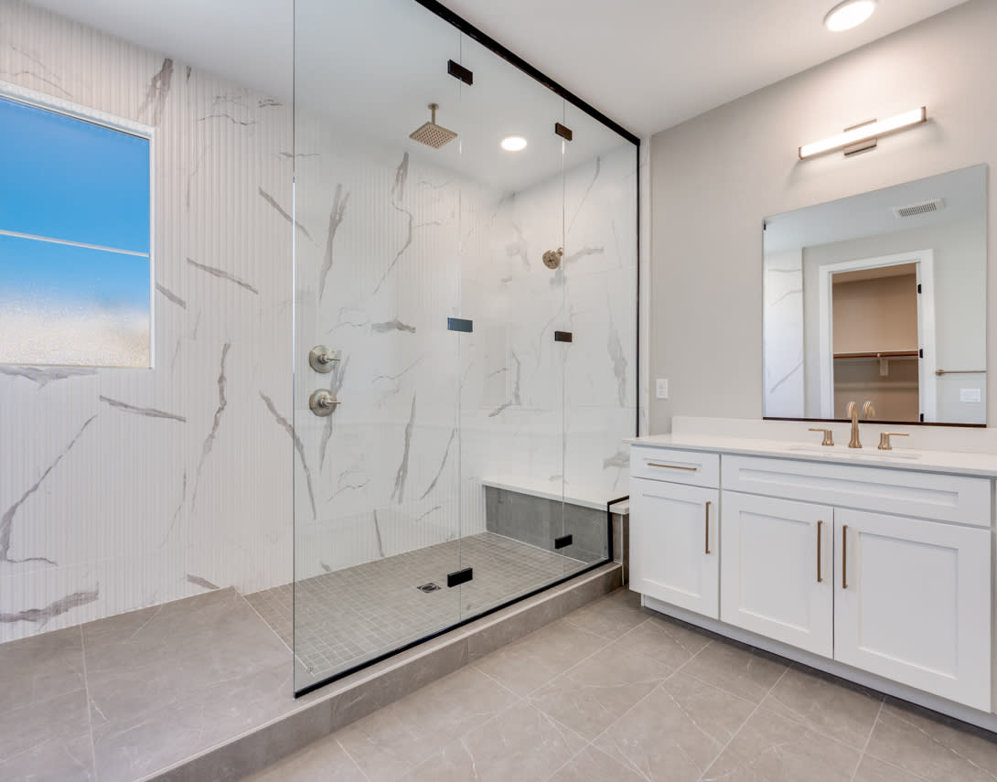 A modern, well-lit bathroom with a large glass shower enclosure, white vanity cabinets, and a view of the sky through a window.