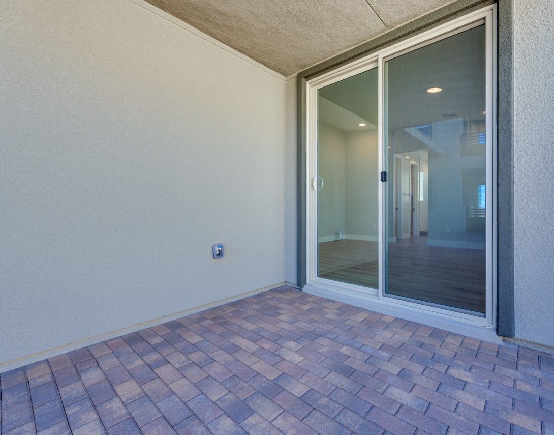 A paved brick floor leads to a sliding glass door, revealing a hallway beyond in a modern, minimalist interior space.