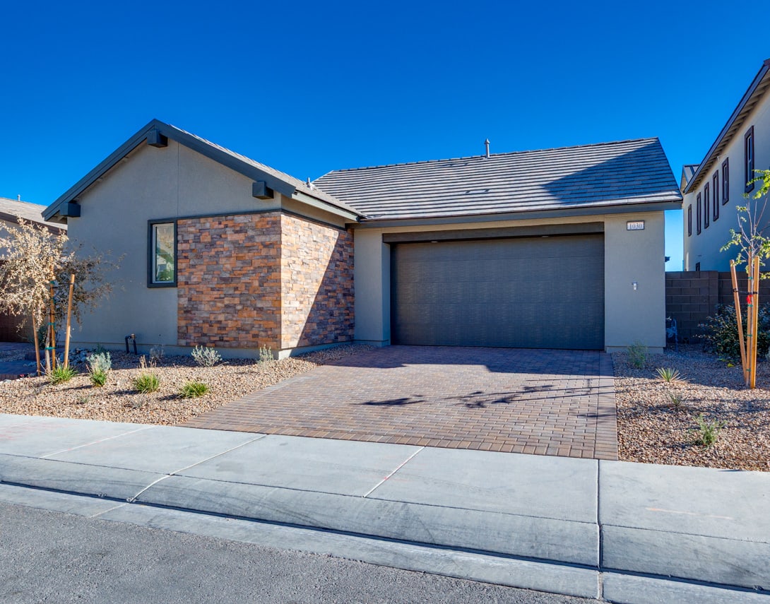 A modern, single-story house with a brick exterior, a garage door, and a paved driveway in the foreground, set against a clear blue sky in the background.