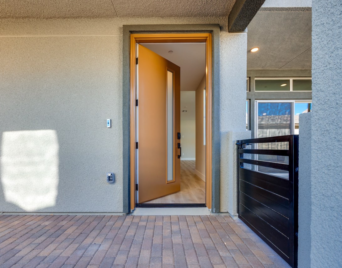 The image shows a modern entryway with a wooden door and a brick patio leading to a hallway with a wooden floor.