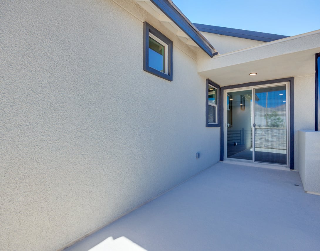 A modern, minimalist exterior with a gray stucco wall, a sliding glass door, and a small window, set against a clear blue sky.