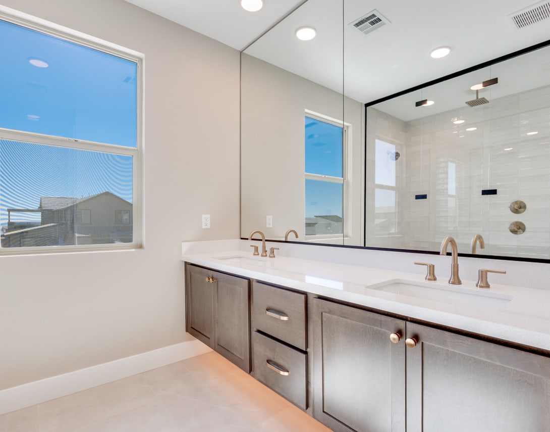 A modern and spacious bathroom with a large vanity, double sinks, and a large mirror, set against a backdrop of windows offering a scenic view.