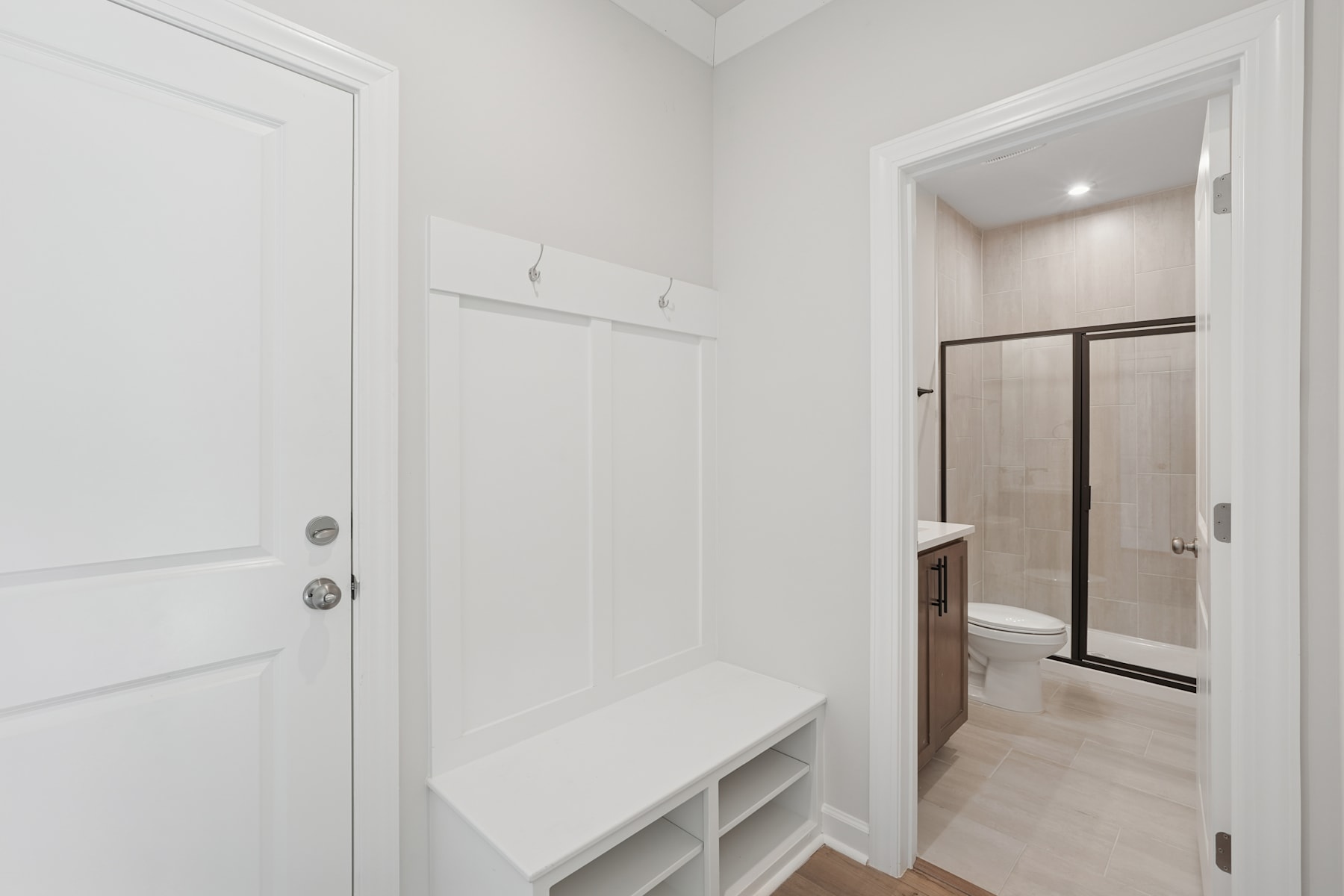 A bright, minimalist entryway with a white door, a built-in bench, and a hallway leading to a bathroom.