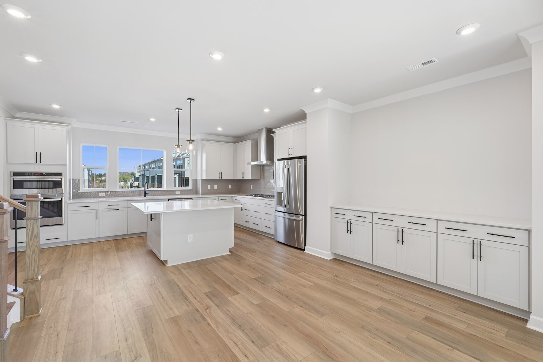 A modern, spacious kitchen with white cabinets, stainless steel appliances, and hardwood floors, with a view of the outdoors through large windows.