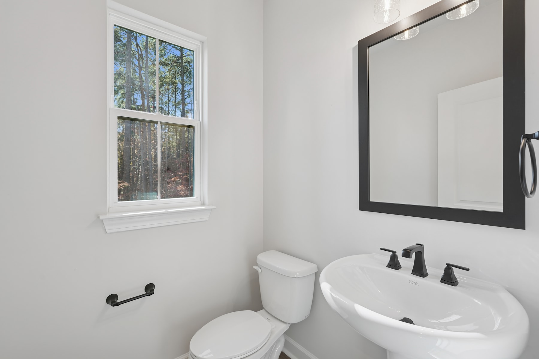 A modern, well-lit bathroom with a white toilet, a pedestal sink, and a large framed mirror on the wall. The room has a clean, minimalist aesthetic with a window overlooking a wooded outdoor area.