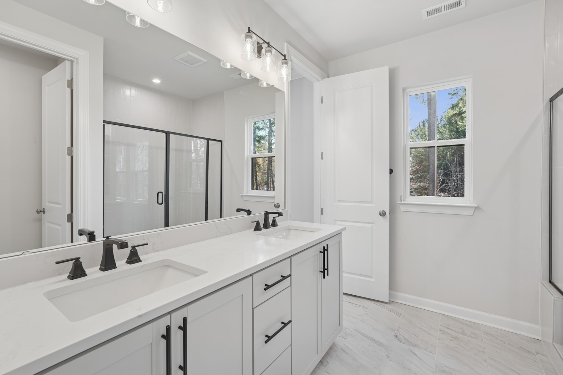 A modern, well-lit bathroom with a double vanity, a large mirror, and a window overlooking the outdoors.