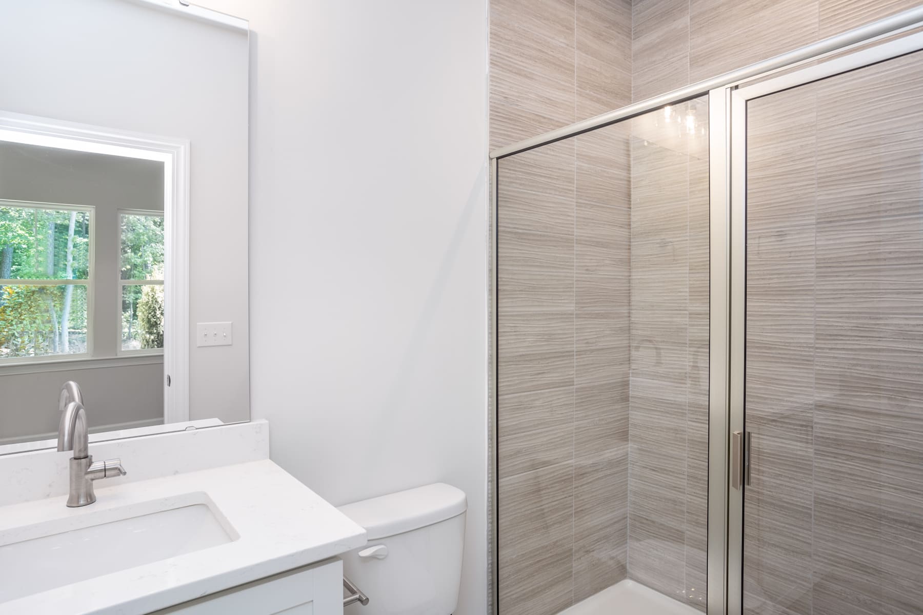 A modern, minimalist bathroom with a white sink, tiled walls, and a glass shower enclosure, overlooking a lush outdoor view through a window.