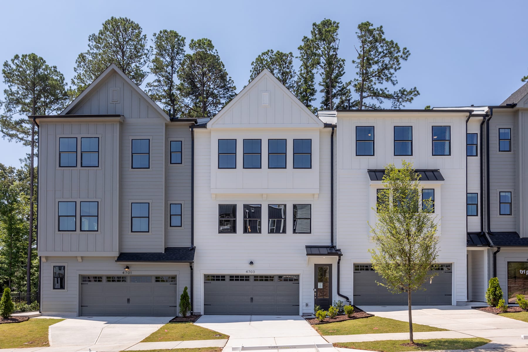 A modern multi-story townhouse complex with gray siding, peaked roofs, and garages in the foreground, surrounded by lush green trees in the background.