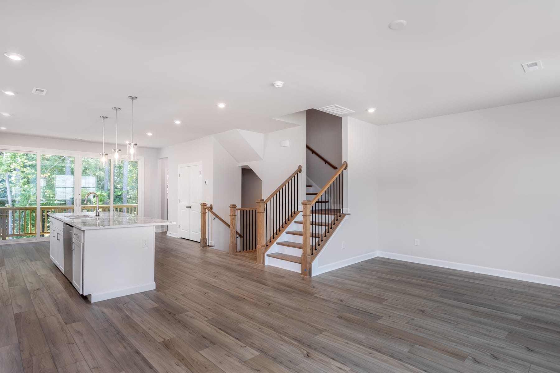 A spacious and bright open-concept interior with a wooden staircase, hardwood floors, and large windows overlooking a lush outdoor environment.
