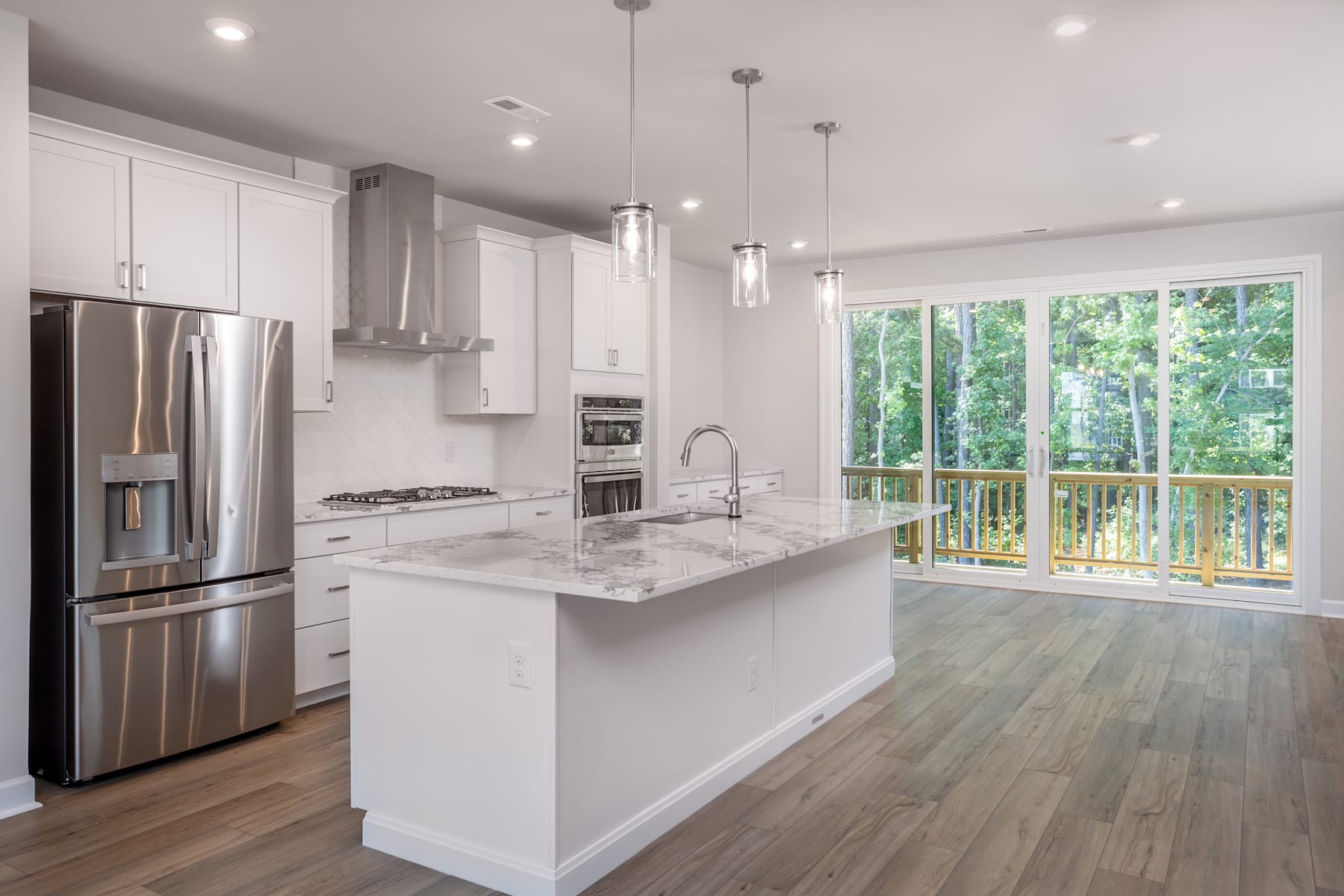 A modern, bright kitchen with white cabinets, stainless steel appliances, and a large window overlooking a wooded outdoor area.