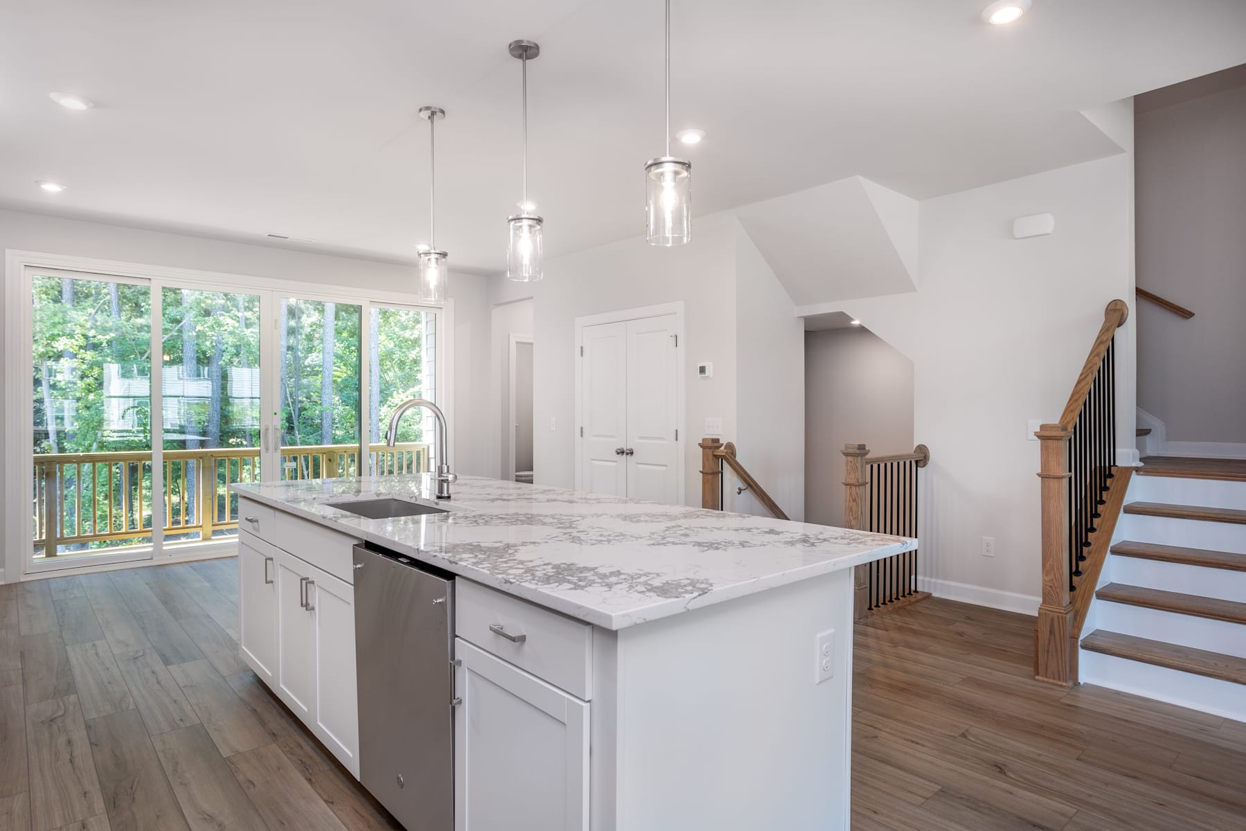 A modern, bright kitchen with white cabinets, marble countertops, and pendant lighting, set against a backdrop of large windows overlooking a lush outdoor area.