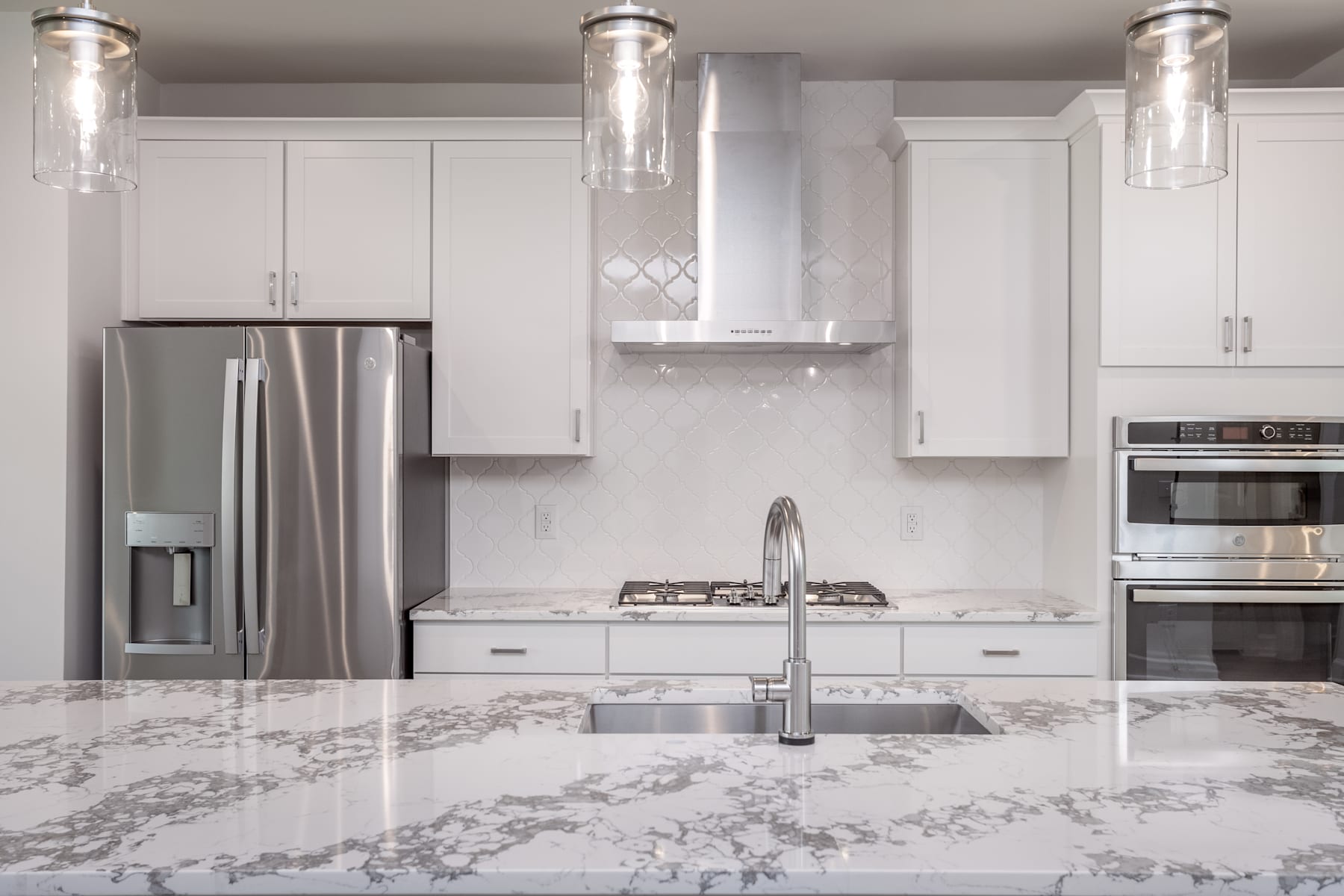 A modern, white kitchen with stainless steel appliances, marble countertops, and pendant lighting fixtures.