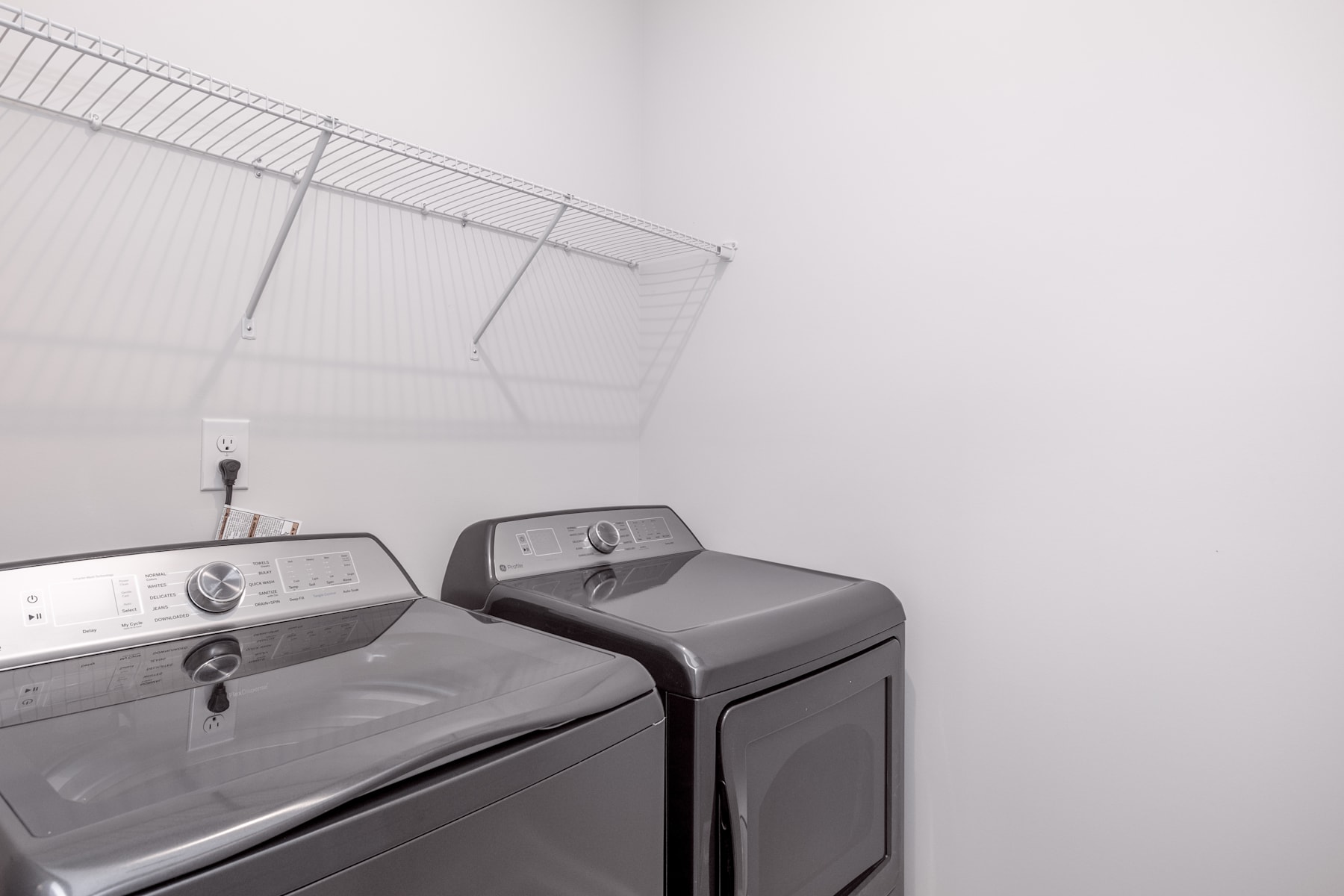 A modern washing machine and dryer set up in a small laundry room, with a wire rack visible in the background.