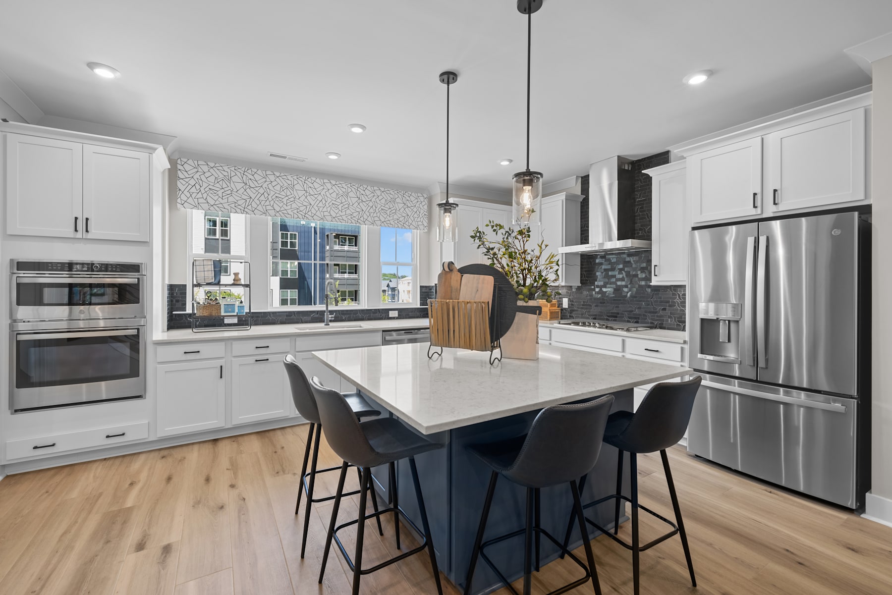 A modern, well-lit kitchen with white cabinets, a large island with bar stools, and stainless steel appliances, set against a backdrop of windows overlooking a cityscape.
