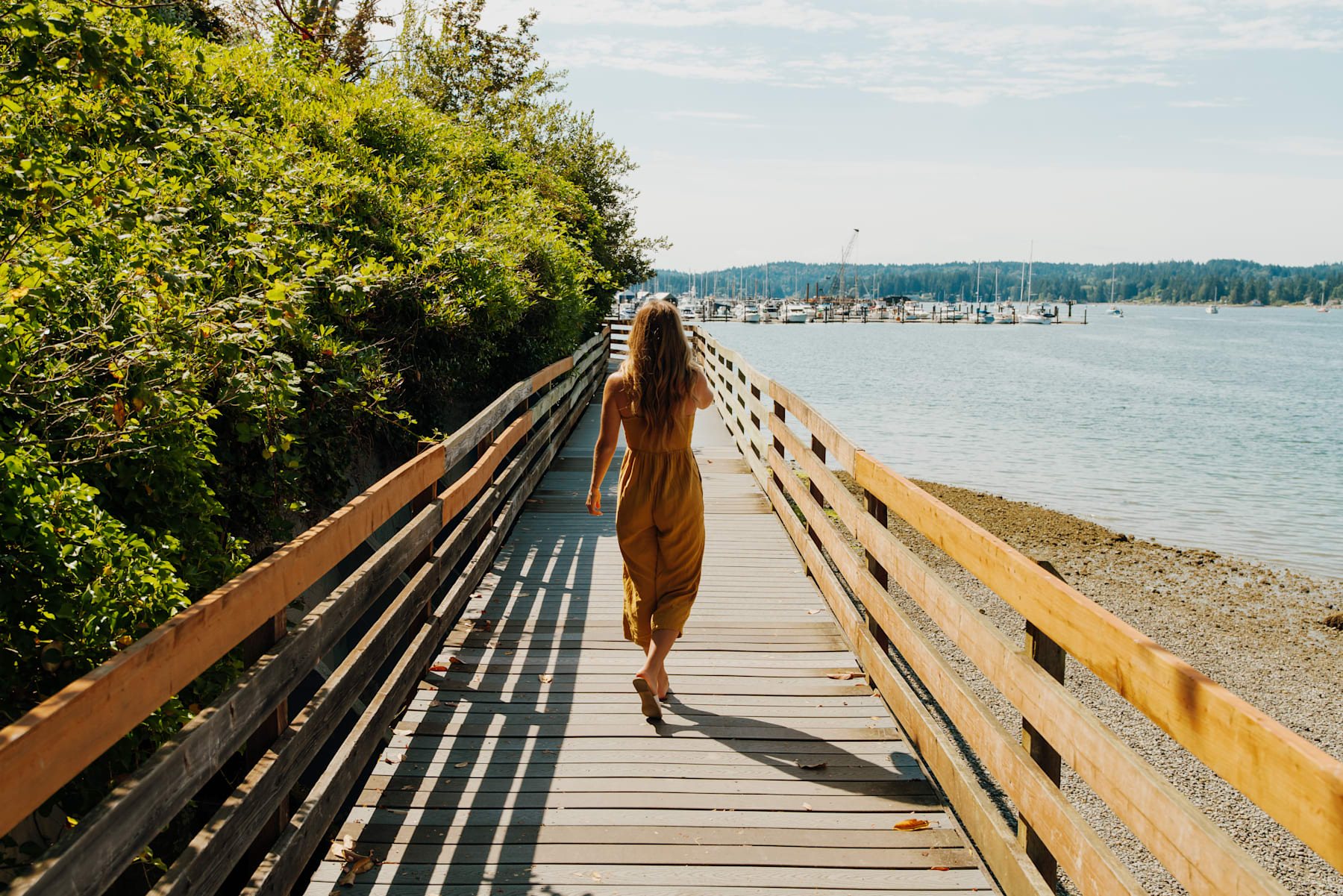A wooden walkway leads through lush greenery towards a scenic body of water, with boats visible in the distance.