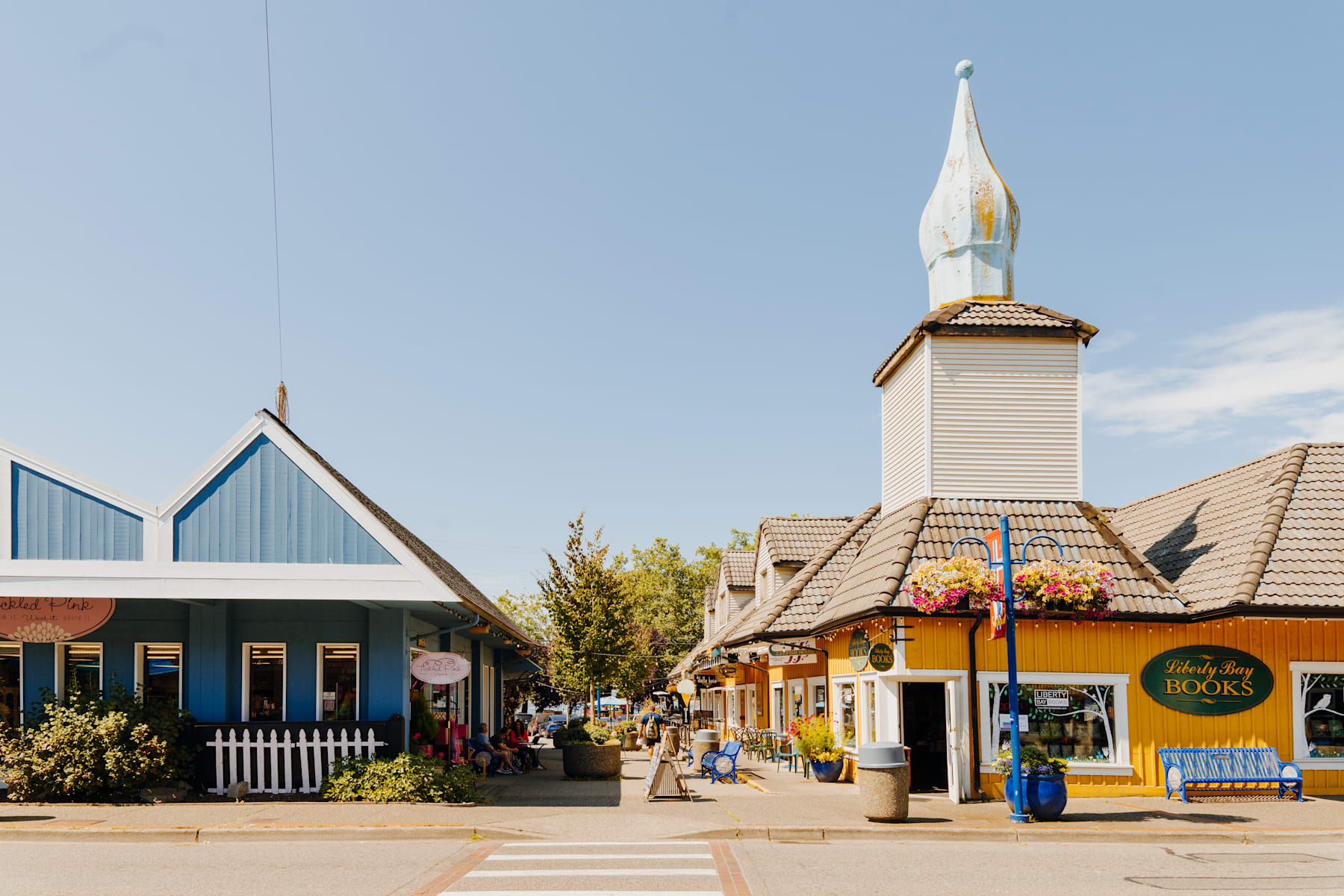 A quaint and colorful village street with a distinctive spire-topped building, surrounded by shops and pedestrians, set against a clear blue sky.