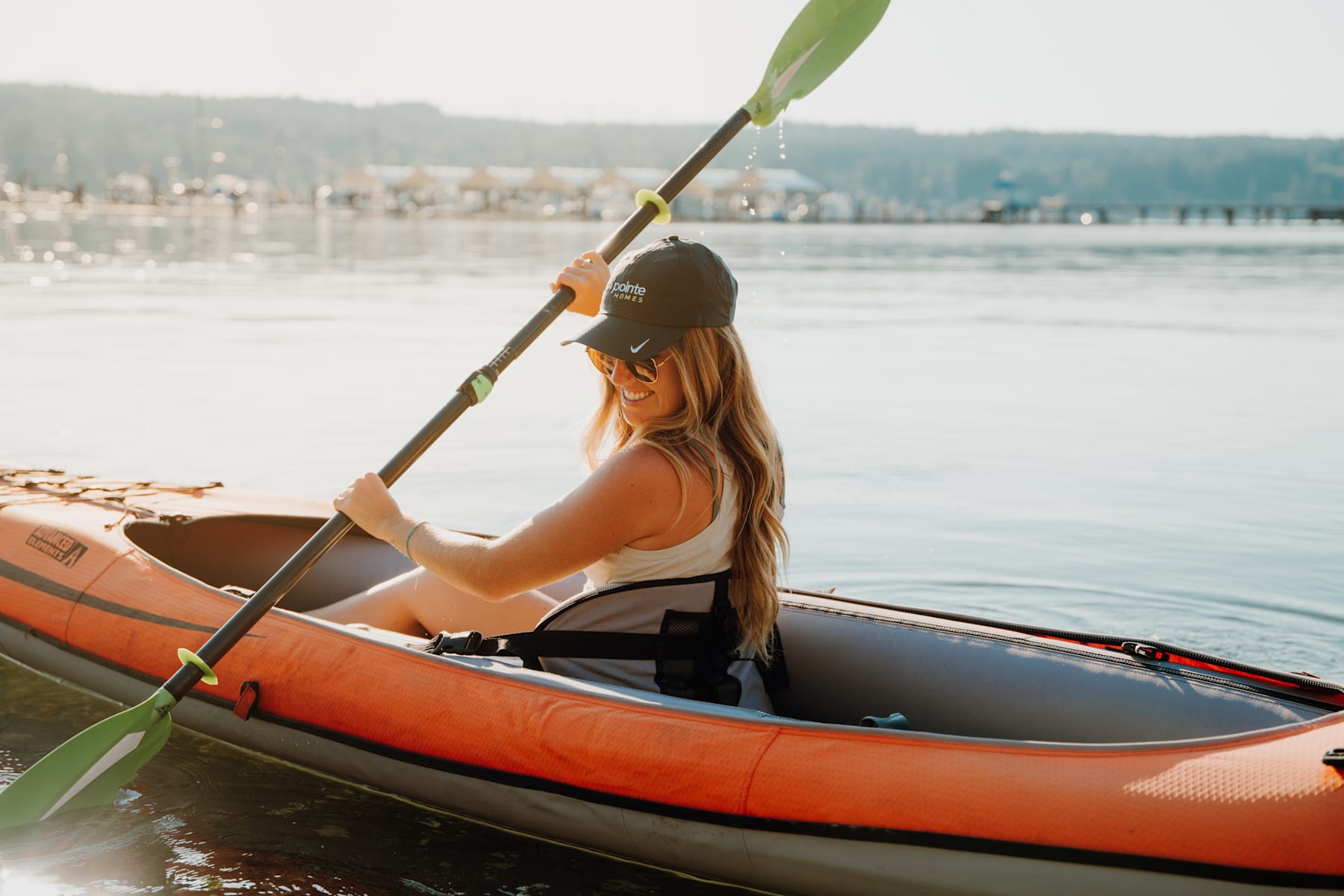 A woman in a bright orange kayak paddles on a serene lake surrounded by mountains in the background.
