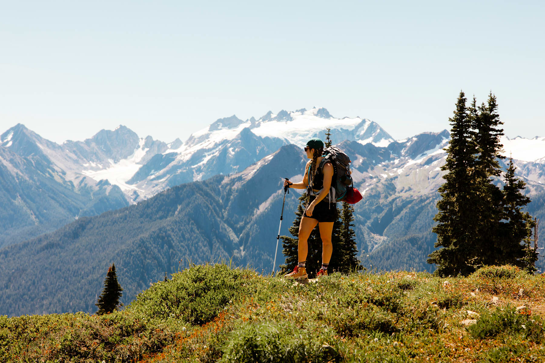 A person in hiking gear stands on a grassy hill, gazing out at the majestic snow-capped mountains in the distance, surrounded by a lush, forested landscape.
