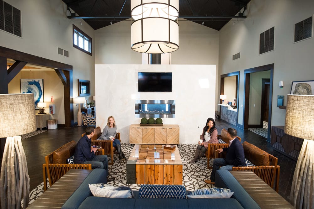 A modern and stylish lobby or lounge area with wooden furniture, patterned rugs, and a large chandelier overhead, with several people seated and conversing.