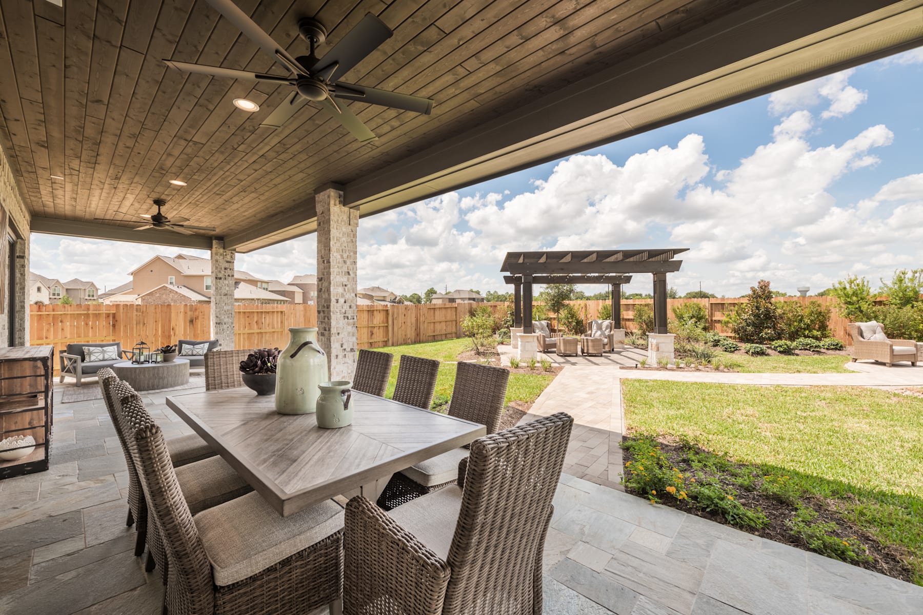 A spacious outdoor patio with a wooden ceiling, ceiling fans, and a large dining table surrounded by wicker chairs, overlooking a well-manicured backyard with a pergola and lush greenery.