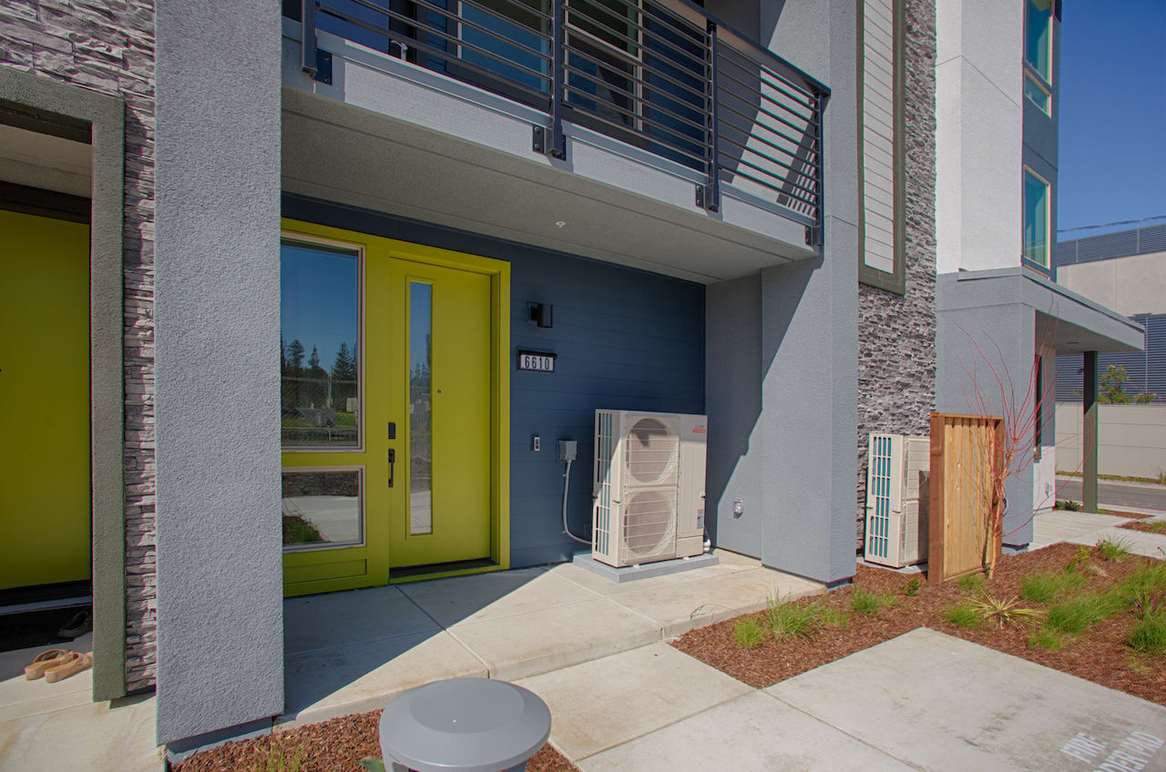 A modern, multi-story building with a yellow front door, a white air conditioning unit, and a concrete walkway leading to the entrance.