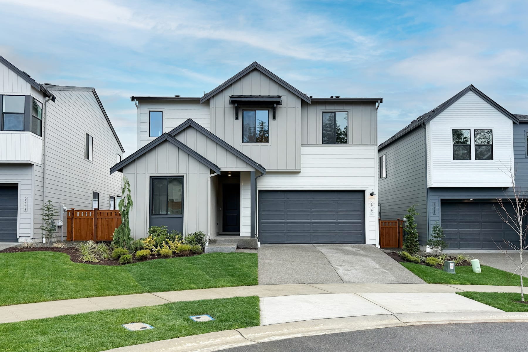 A modern, two-story residential house with a well-manicured lawn and landscaping in the foreground, set against a backdrop of a clear blue sky with scattered clouds.