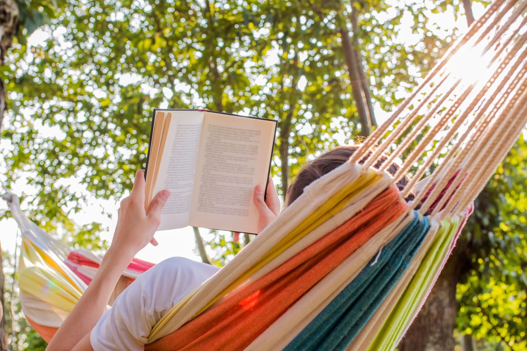 A person is relaxing in a colorful hammock, reading a book amidst the lush greenery of the surrounding trees.