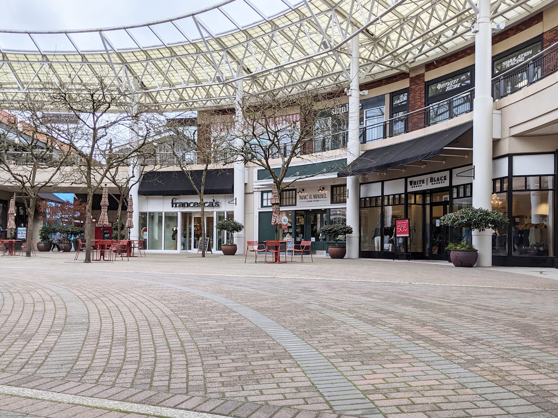 A modern outdoor shopping mall with a glass-enclosed atrium, brick-paved walkways, and various retail stores visible in the background.