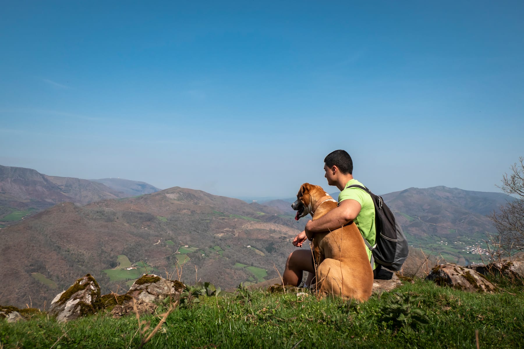 A person wearing a backpack stands on a grassy hill, overlooking a mountainous landscape in the background.