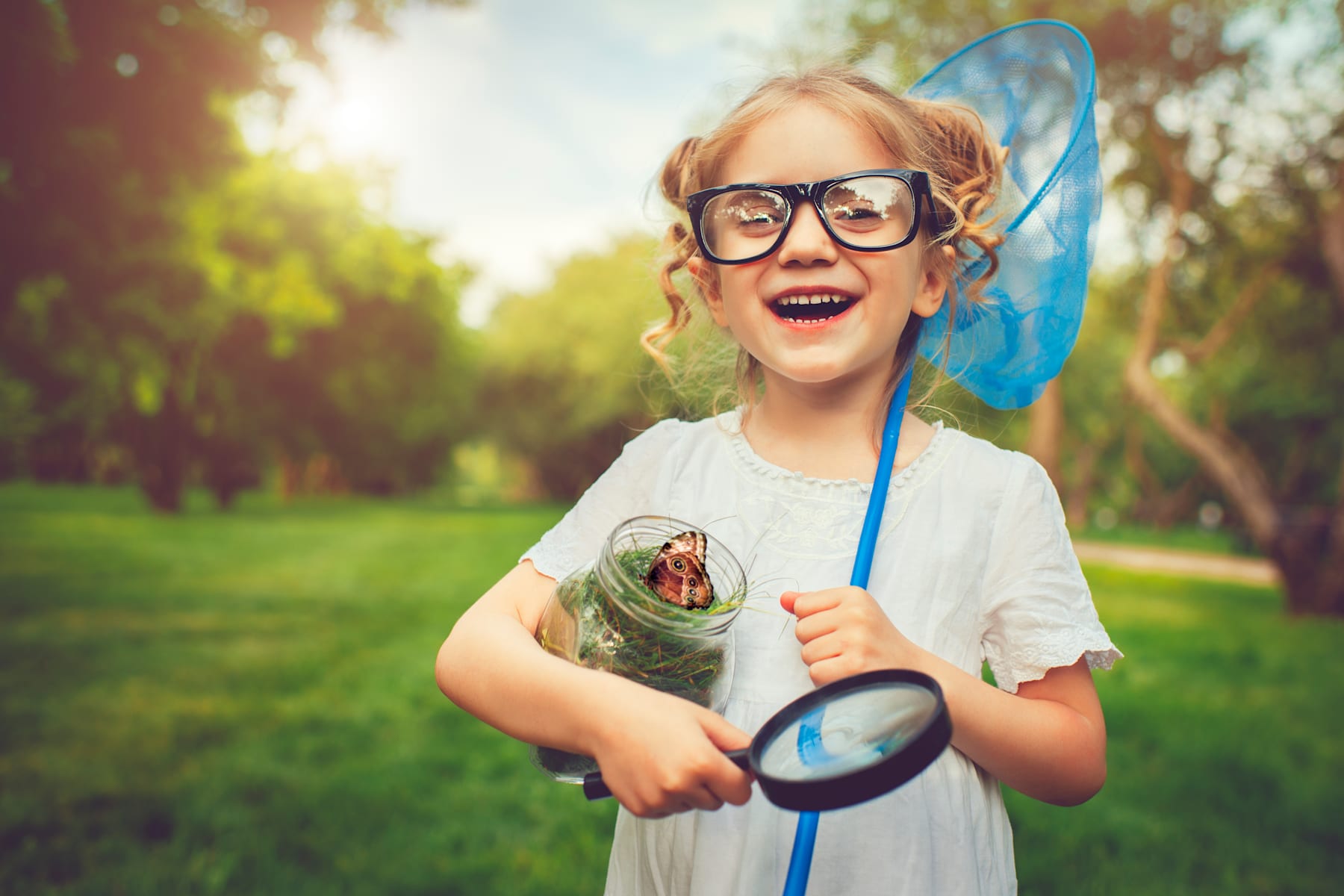 A young girl with curly blonde hair and glasses is holding a magnifying glass and a butterfly net, standing in a lush green park with trees in the background.