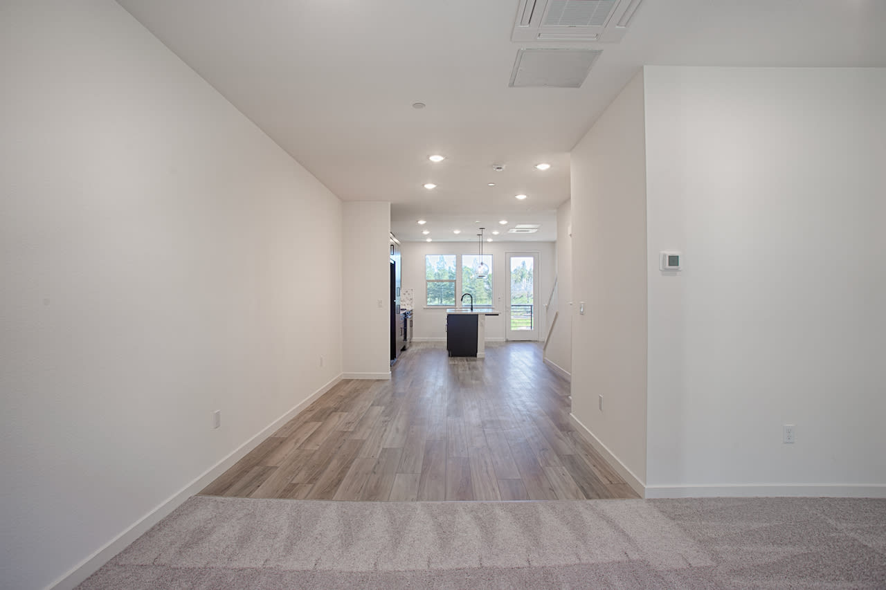A bright, open hallway with white walls, a hardwood floor, and recessed lighting leading to a glass door that provides a view of the outdoors.