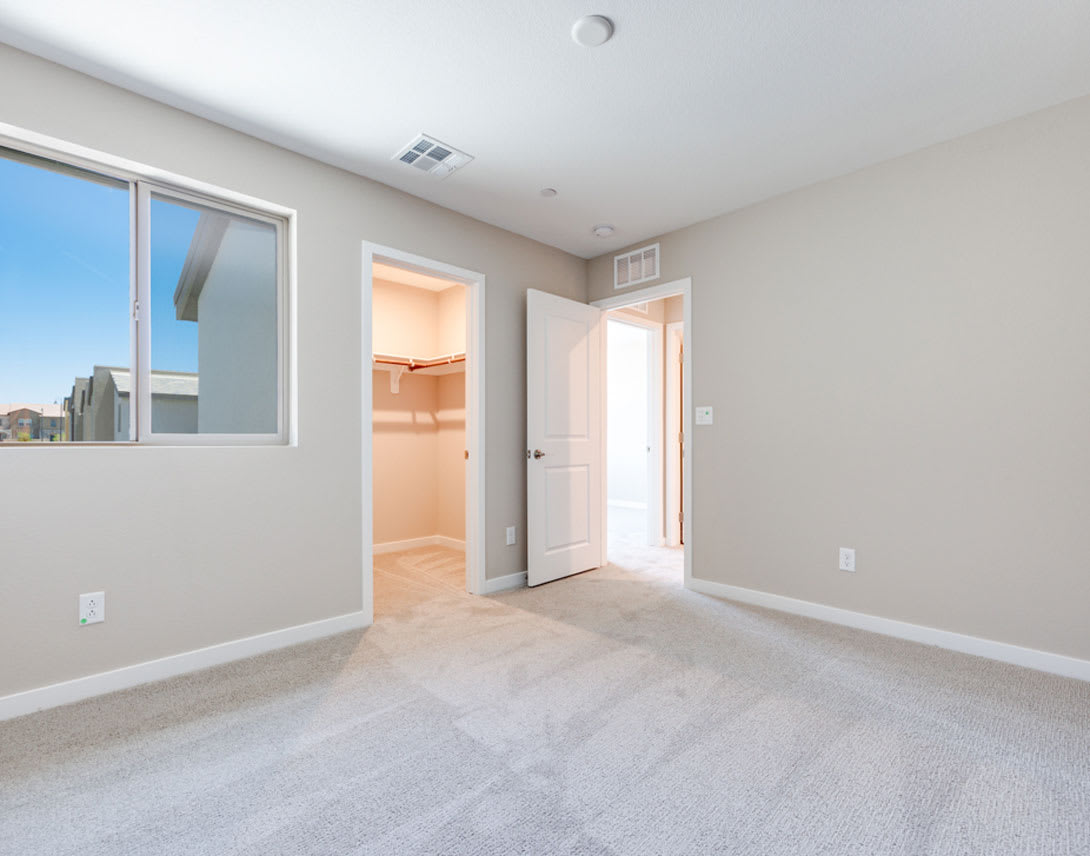 A spacious, well-lit room with a large window, a closet, and a door leading to another room, all set against a neutral color palette and carpeted flooring.