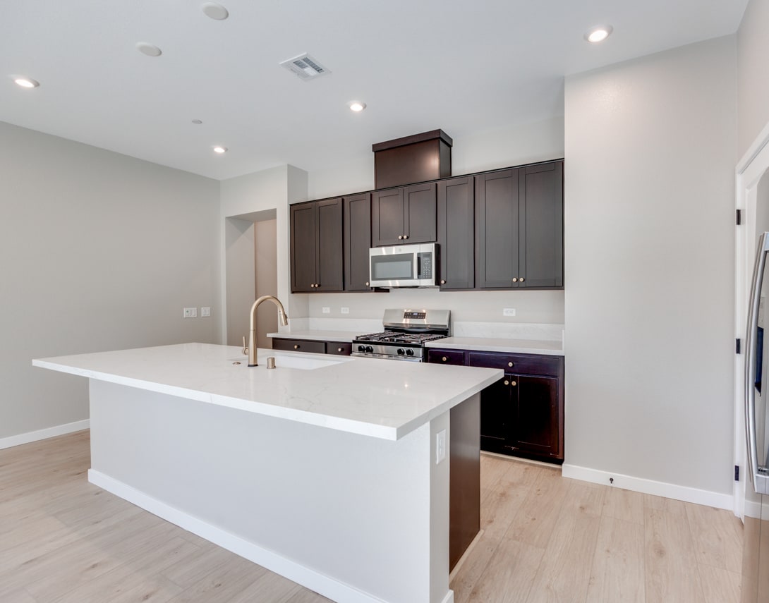 A modern, open-concept kitchen with dark wood cabinets, white countertops, and stainless steel appliances, set against a light-colored wall and hardwood flooring.