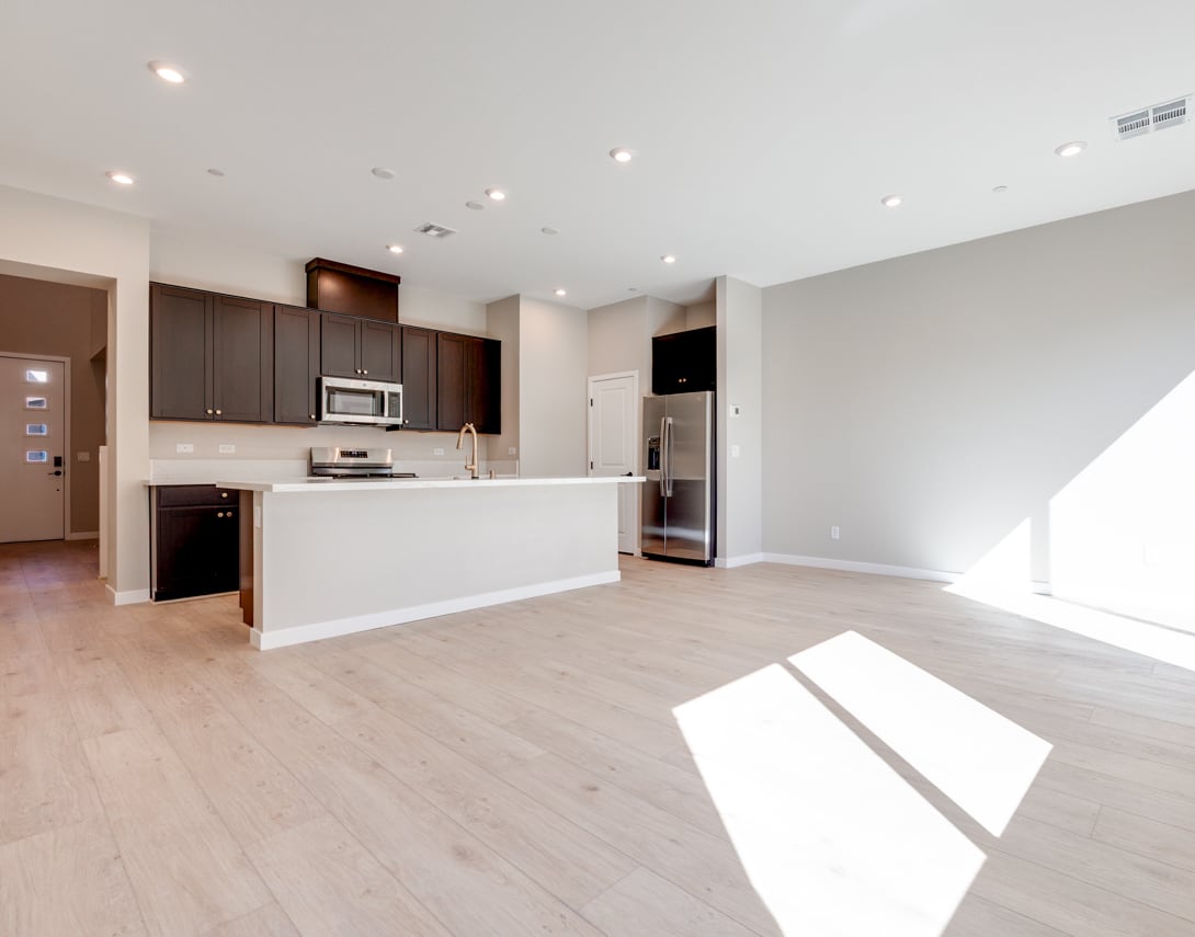 A modern, open-concept kitchen and living space with dark wood cabinets, stainless steel appliances, and light hardwood floors, bathed in natural sunlight.