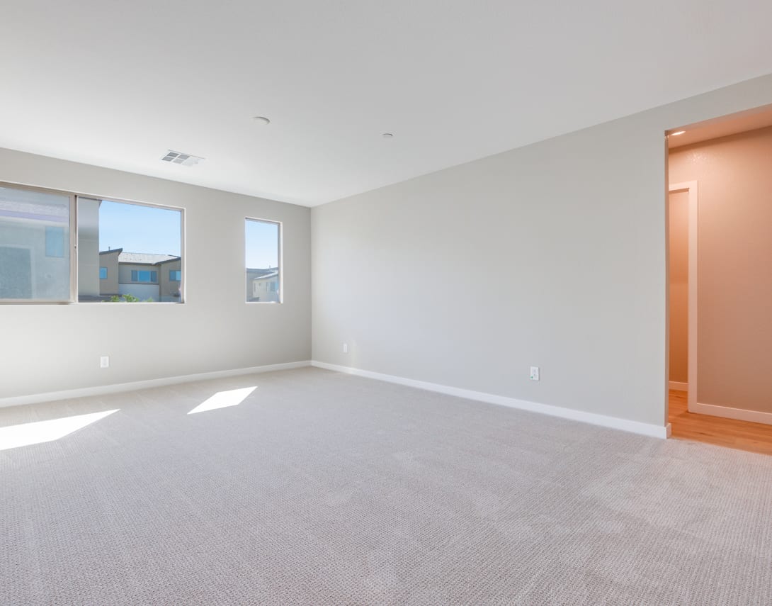 An empty, spacious room with white walls, a carpeted floor, and large windows allowing natural light to fill the space.