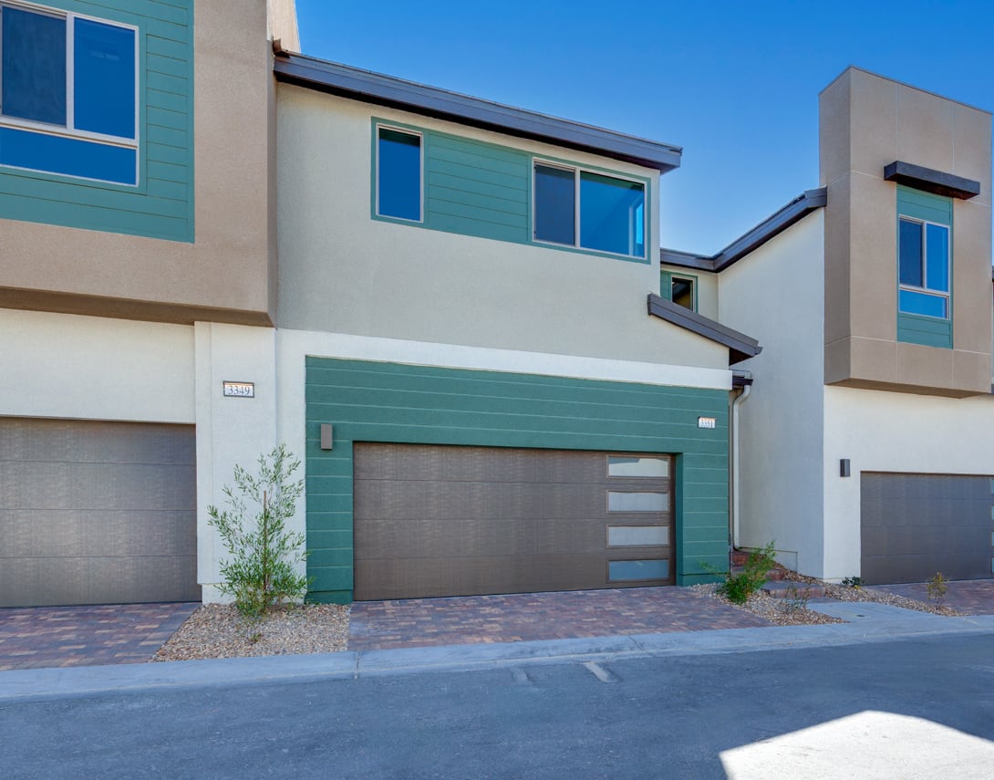 The image depicts a modern, multi-story residential building with a mix of colors and materials, including gray, blue, and green tones. The foreground features a paved driveway leading to a garage door, and the background shows a clear blue sky.