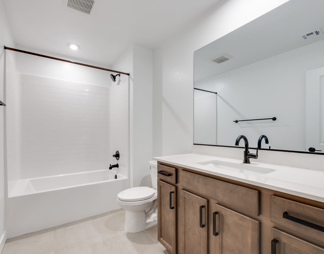 A modern bathroom with a white tiled wall, a wooden vanity with a white countertop, and a toilet in the background.