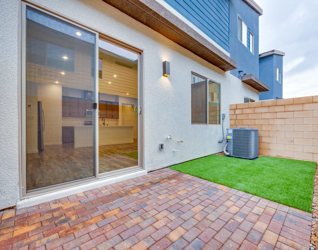 A modern, two-story residential building with a paved brick patio, a small grassy area, and large glass windows in the foreground.