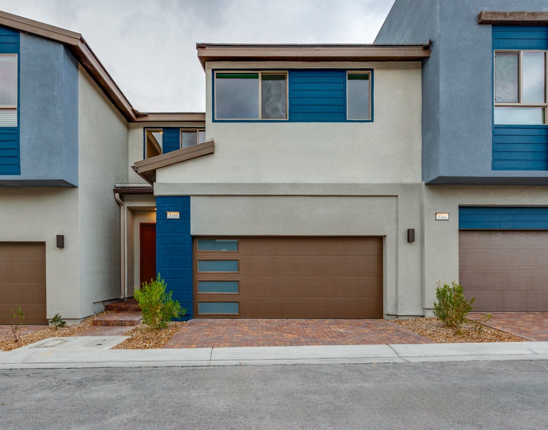 A modern, multi-story townhouse with a garage door, blue accents, and a paved driveway in the foreground, set against a cloudy sky backdrop.
